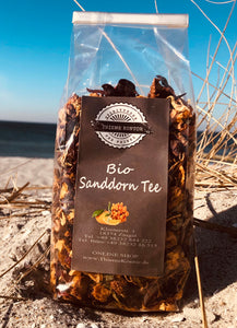 Bild zeigt eine Großaufnahme einer 100 Gramm Packung Bio Sanddorn Tee von Thieme Kontor am Strand von Zingst. Im Hintergrund sieht man die Ostsee.